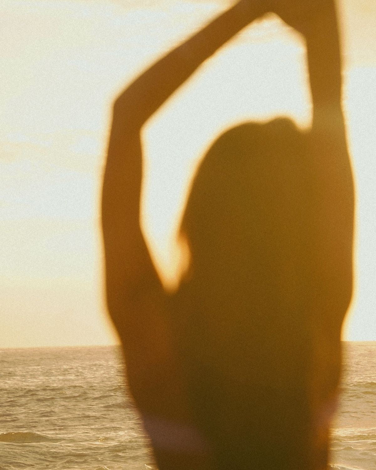 Woman seen from behind, arms raised overhead, facing the sea in golden light