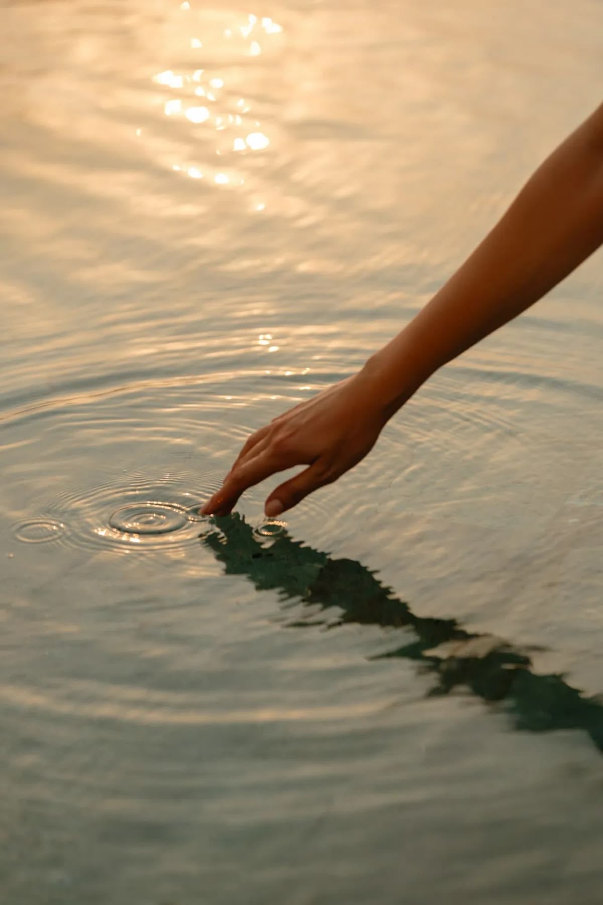 A hand touching calm water, ripples spreading in golden light